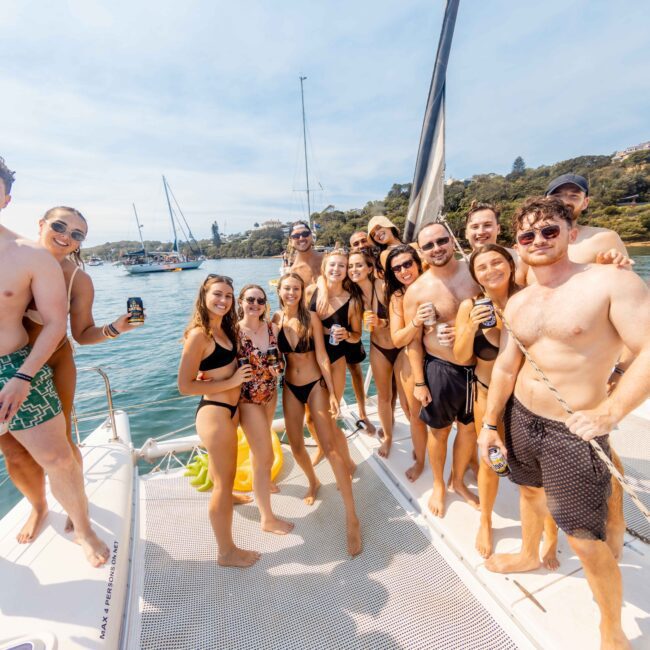 A group of people in swimwear are posing and smiling on a boat. They are holding drinks and enjoying a sunny day on the water. Other boats and a scenic shoreline are visible in the background.