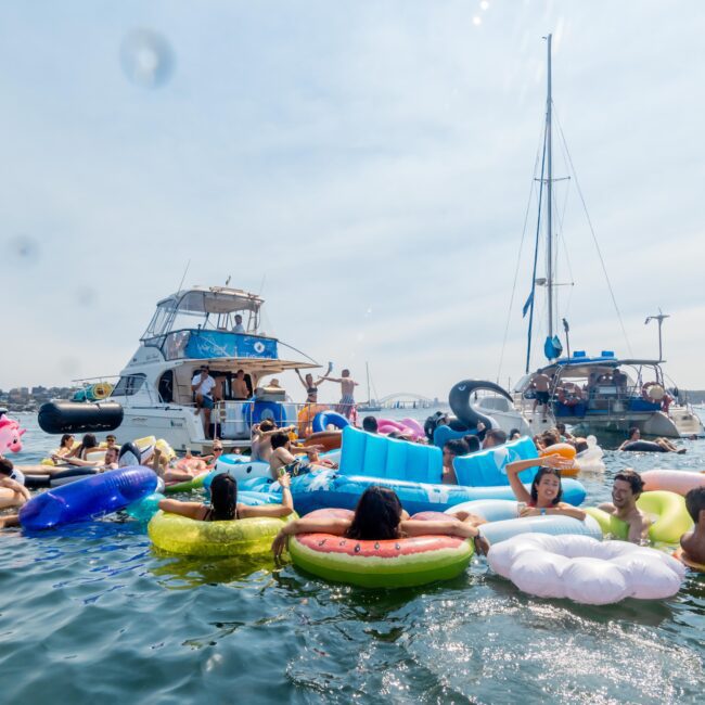 People enjoying a sunny day on inflatable rafts in a large body of water, surrounded by boats. The scene is lively with colorful floats and people swimming, socializing, and relaxing under a clear blue sky.