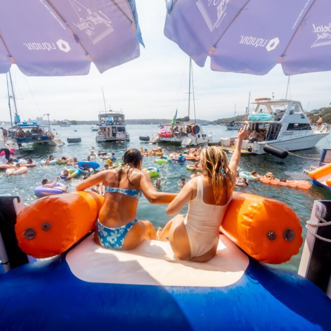 Two people sit on a large inflatable with orange sides, looking out at a crowded scene of people on floaties in the water. Several boats are anchored nearby. Blue canopy shades part of the image. Festive atmosphere.