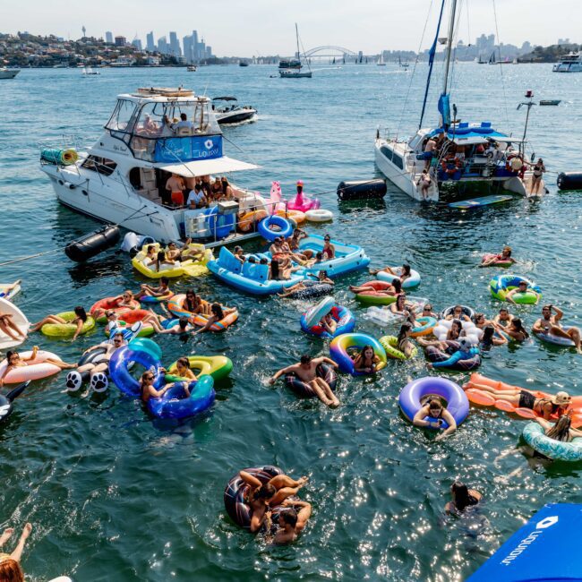 A crowded scene of people enjoying a sunny day on the water. They are floating on colorful inflatables near several boats, with a cityscape in the background. The atmosphere is lively and festive.