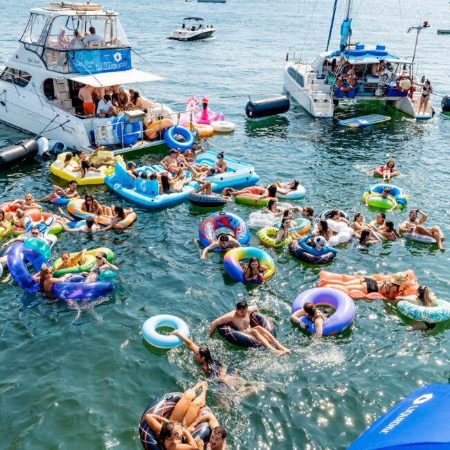 A group of people relax on colorful inflatable rings and floaties in a sunny, busy bay. Several boats are anchored nearby, and the city skyline with a bridge is visible in the background. The scene is lively and vibrant.