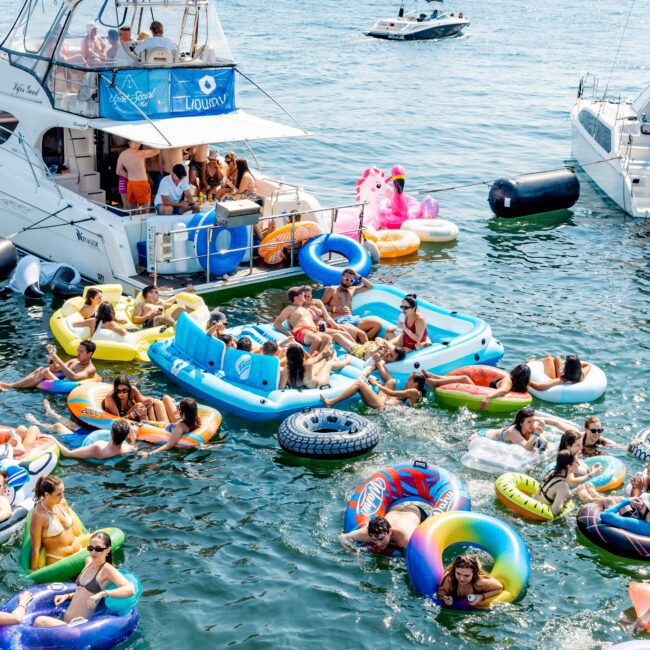 A vibrant scene of people enjoying a sunny day on a harbor, relaxing on colorful inflatable floats and swimming near boats. The city skyline and a bridge are visible in the background against a clear blue sky.