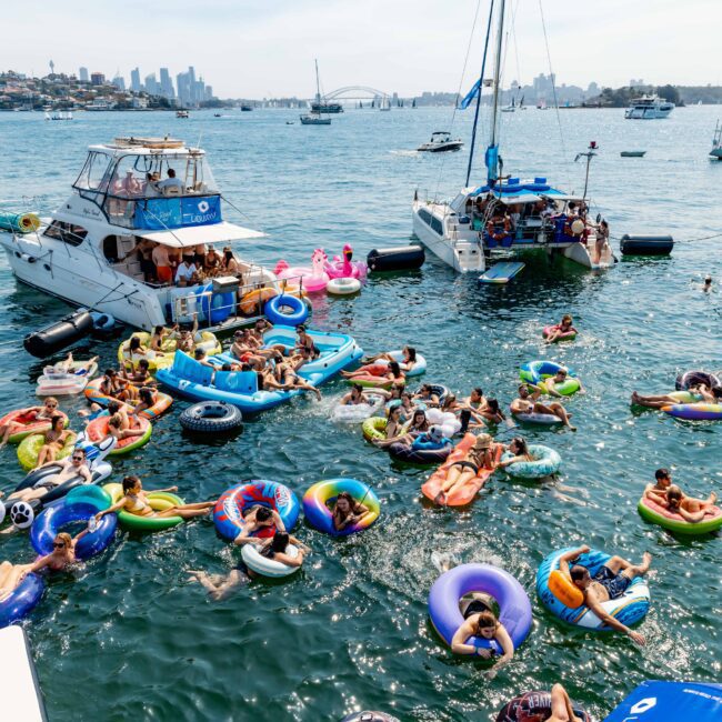 A large group of people enjoying the water on colorful inflatable floats near several boats. The background shows a city skyline. It's a sunny day, and the scene is vibrant and lively.
