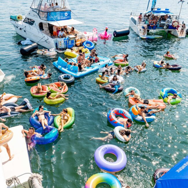 People enjoying a sunny day on a body of water, floating on colorful inflatable tubes and rafts. Boats are anchored nearby and the background shows a city skyline. The atmosphere is lively with people swimming and relaxing.