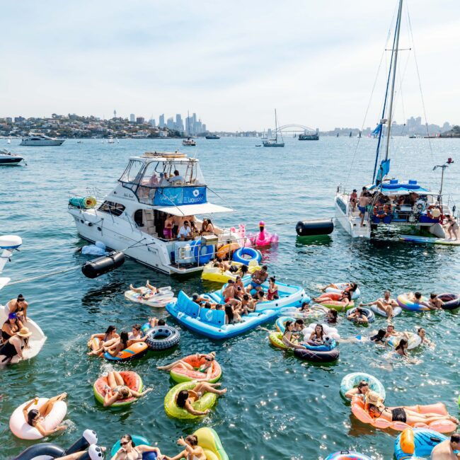 A lively scene on the water with numerous people enjoying a sunny day on various colorful inflatables. Boats are anchored nearby, and the distant city skyline can be seen under a clear blue sky.