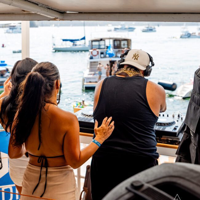 A woman stands next to a DJ on a yacht, placing her hand on his back. They are overlooking a scene filled with boats on the water. The setting is lively, with a bright, sunny backdrop. The Yacht Social Club logo is visible in the corner.