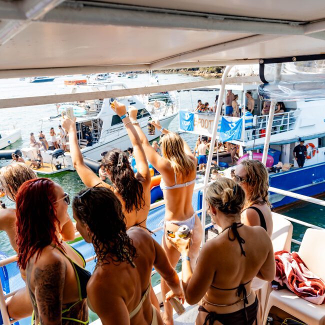 A group of people in swimsuits are on a boat, some with their hands raised. Other boats are visible in the background. The weather is sunny, and the atmosphere appears lively and festive.