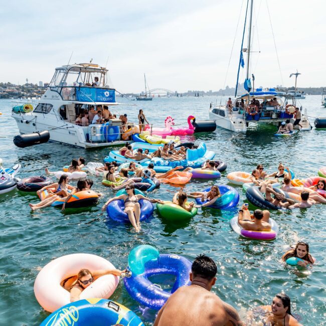 A lively group of people enjoying a sunny day on the water, lounging on colorful inflatable floats. Two boats are anchored nearby. The scene is festive, with music, relaxed sunbathers, and swimmers in a large, open body of water.