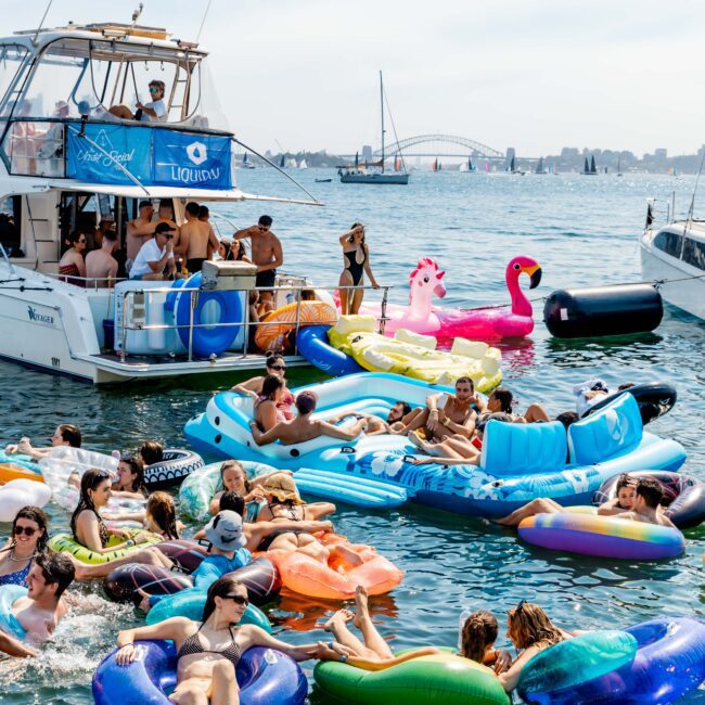 A vibrant scene of people relaxing on colorful inflatables in the water near boats on a sunny day. The background features a bridge and numerous yachts. The atmosphere is festive and lively, with music playing from a nearby boat.