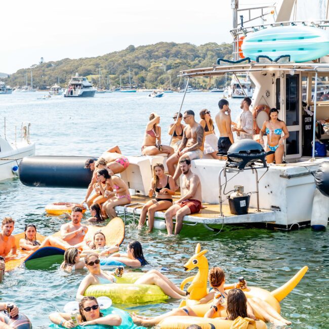 People enjoying a sunny day at a lake with inflatable floats, including a kangaroo and pineapple. A boat is docked nearby with more people sitting on it. Other boats and forested land are visible in the background.