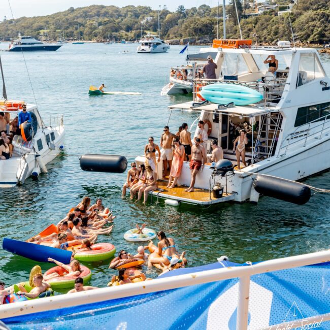 A large group of people enjoying a party on boats, floating devices, and paddleboards in a sunny harbor. Several yachts and trees are visible in the background. Decorations and inflatable toys add a festive atmosphere.