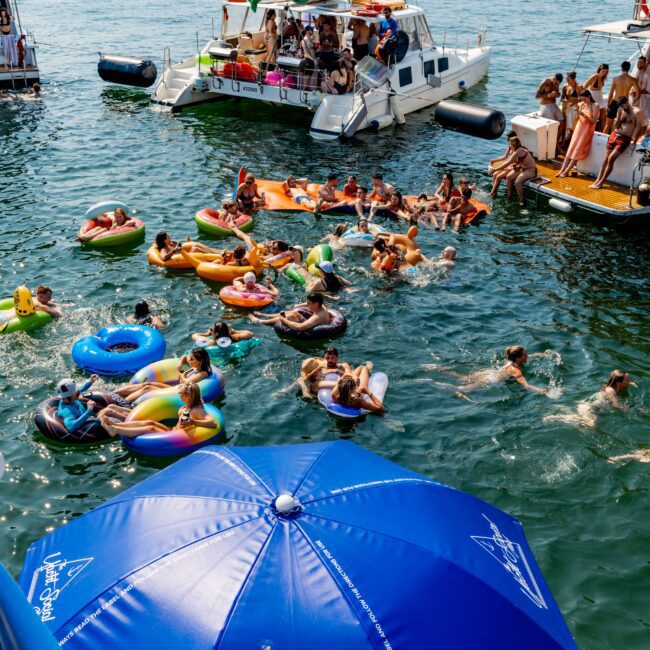 A lively scene on a sunny day with people relaxing on colorful inflatables and swimming in the sea. Several yachts are anchored nearby, and a blue umbrella from The Yacht Social Club is in the foreground. A festive, social gathering atmosphere.