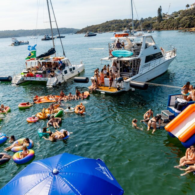 People enjoying a sunny day on the water with boats, inflatables, and a floating slide. Some are swimming, while others relax on yachts. A lively, festive atmosphere is present, with a backdrop of trees, houses, and clear blue skies.