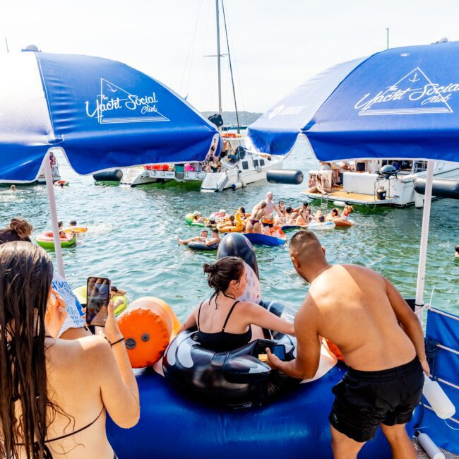 People enjoying a sunny day on the water. Some are relaxing on inflatable floats while others are on a boat nearby. Blue umbrellas provide shade, and a few individuals are taking photos.