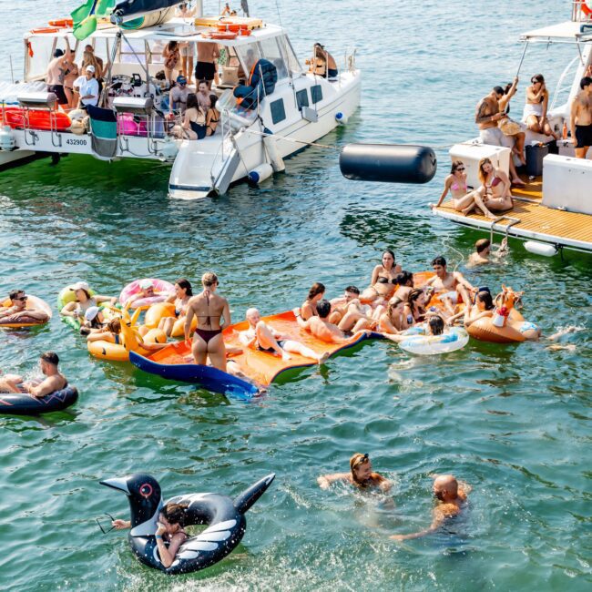 A lively scene with people sunbathing on a floating mat and inflatable toys in the water near anchored boats. There are various boats in the background and a large blue umbrella partially visible in the foreground.