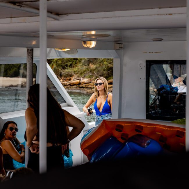 A woman in a blue swimsuit stands on a boat, looking out at the water. Other people relax onboard, and inflatable floaties are visible. The setting appears to be sunny and tropical, with greenery in the background.