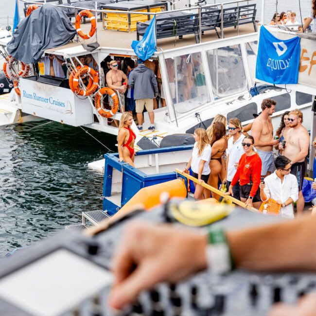 A group of people enjoy a party on two docked boats in a scenic waterfront location. The focus is on a DJ mixing music at a console in the foreground. There are lifebuoys, blue flags, and festive decorations on the boats.