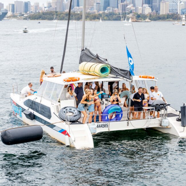 A group of people enjoying a boat party on a catamaran with a city skyline in the background. The boat has a blue flag and inflatable decorations. Some people are standing, and others are sitting, enjoying the cruise on a cloudy day.