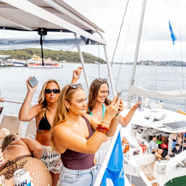 A group of people enjoying a boat party on the water. Some are holding phones, and others have drinks. The boat has a blue flag, with a scenic view of water and land in the background. The atmosphere is lively and festive.