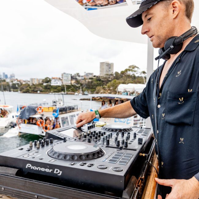 A DJ wearing a black shirt and cap is mixing music on a Pioneer DJ controller on a boat. Behind him, there are other boats and a waterway, with buildings and trees in the background. He has headphones around his neck.