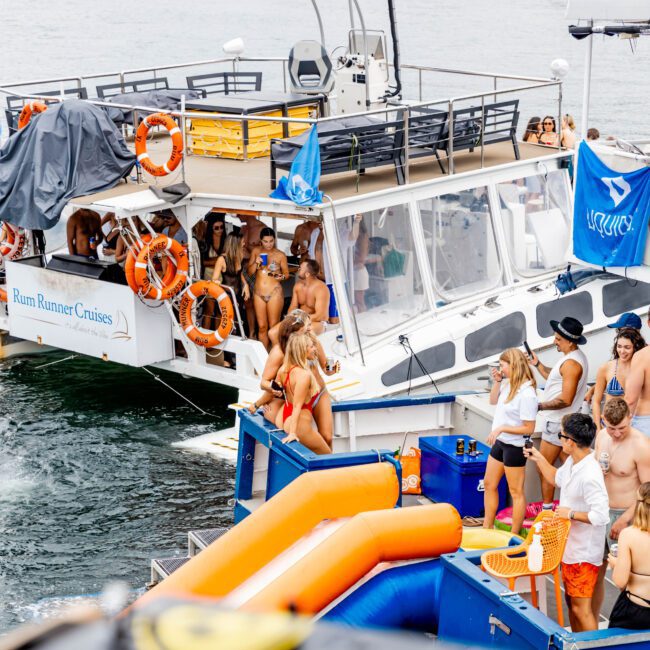 A lively party is happening on two docked boats. People in swimwear are socializing and relaxing. A group is gathered near an inflatable slide on one boat, and others are on the deck of the "Rum Runner Cruises" boat. Flags and life rings are visible.
