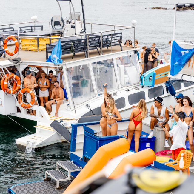 A boat party with people in swimwear enjoying music and dancing. Two women in bikinis are in the foreground, one waving her arm. The boat is equipped with a slide and life vests. Other boats and a shoreline are visible in the background.