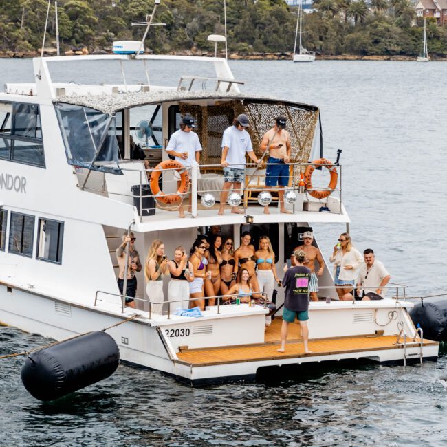 A group of people in swimwear and casual clothing enjoy a party on a yacht with a seated upper deck on a waterway. A bridge is visible in the background, along with lush greenery on the shoreline.
