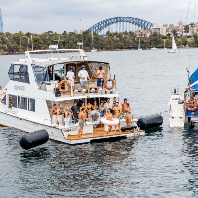 A group of people enjoying a party on a yacht named "Kondor" in a harbor. The scene includes a large bridge and cityscape in the background. Lifebuoys are visible on the boat's railings. The weather appears cloudy.