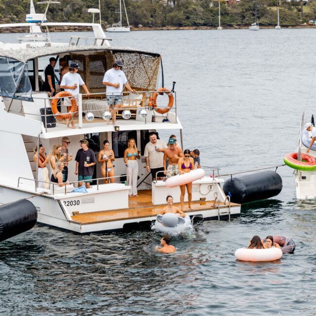 A group of people enjoying a sunny day on a yacht, some swimming in the water with inflatable rings. The Sydney Harbour Bridge is visible in the background. The yacht is anchored, and more people are on the deck observing.