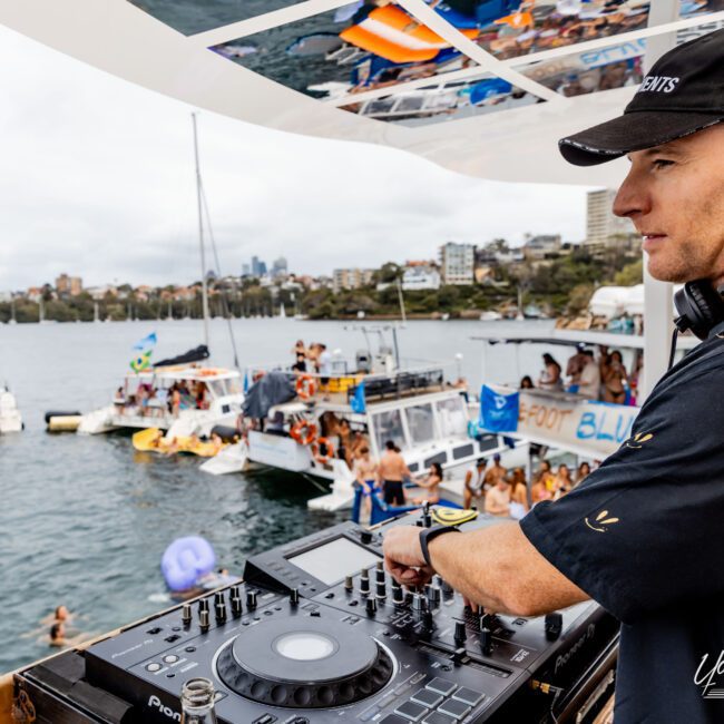 A DJ wearing headphones plays music on a boat, with DJ equipment in front of him. In the background, several boats are gathered on the water, and people are socializing and enjoying the scene. The sky is overcast, and a city is visible in the distance.