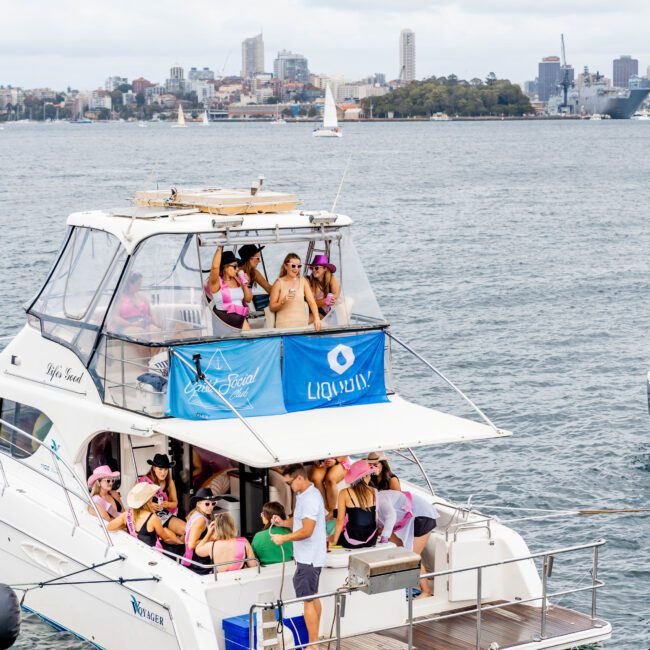 A group of people socialize and dance on a docked yacht named "Infinity." They are gathered around the upper deck, with city skyscrapers and a sailboat visible in the background under a cloudy sky. The yacht displays "Yacht Social" and "Liquid" banners.