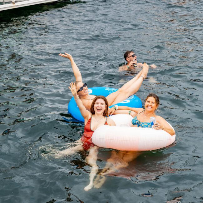 Four people are enjoying the water near some boats. Two people are floating on inner tubes, smiling, and waving, while two others swim nearby, relaxing and smiling in the water.