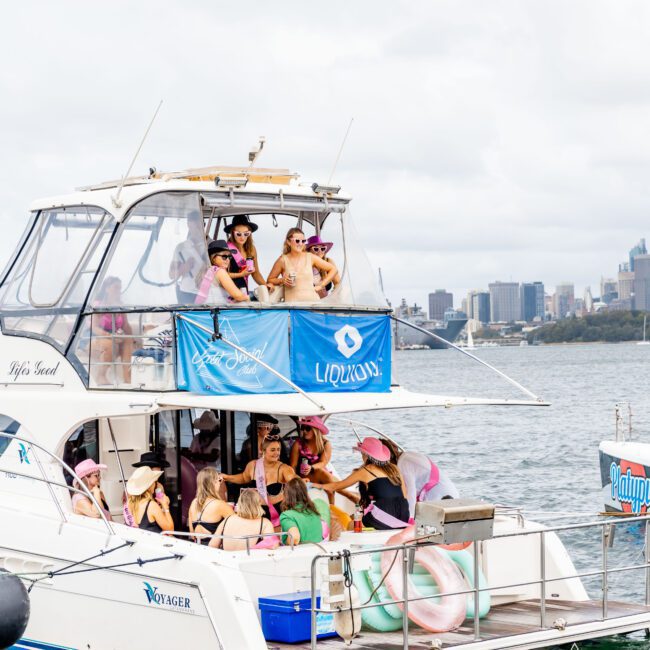 A group of people in festive attire enjoy a party on a white yacht named "Voyager" with a "Liquidn" banner. They're wearing hats and posing cheerfully. The boat is on water with a city skyline visible in the background under a cloudy sky.