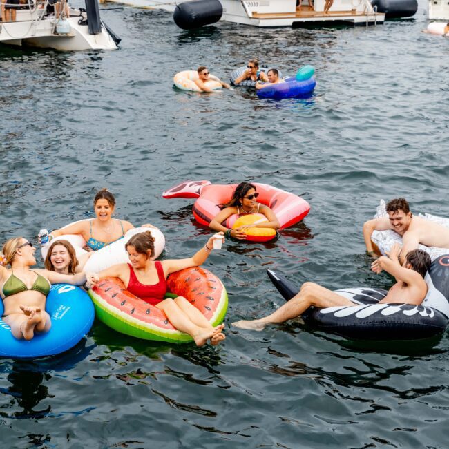 A group of people enjoying a day on the water, lounging on various inflatable floats. The floats include a blue ring, red float, watermelon slice, swan, and a boat-shaped float. Some are holding drinks and smiling, with boats visible in the background.