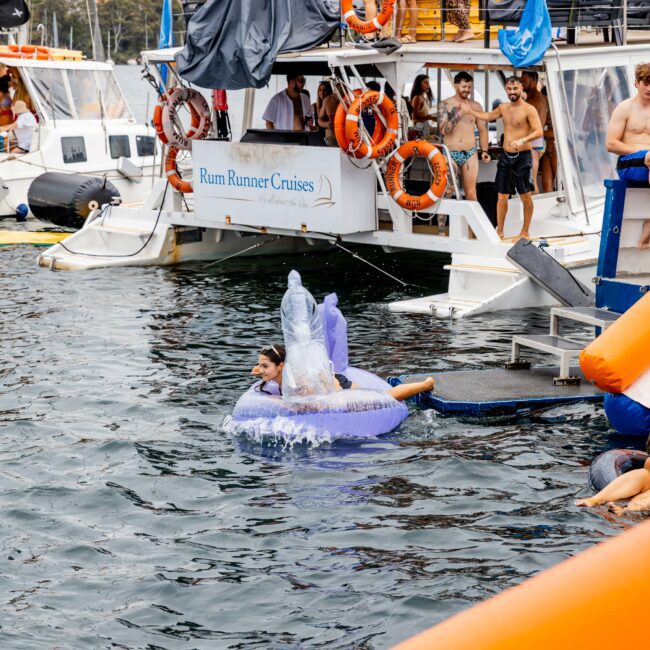 People on a boat party; some are wearing swimsuits and enjoying drinks. A person is riding an inflatable unicorn in the water, with splashing motion. Lifebuoys and a sign reading "Rum Runner Cruises" are visible on the boat. City skyline in background.