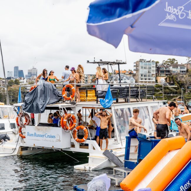 A lively gathering on a boat named "Rum Runner Cruises" docked by the waterfront. People enjoy the day with inflatable toys, including a swan. Buildings are visible in the background, and umbrellas partially frame the scene.