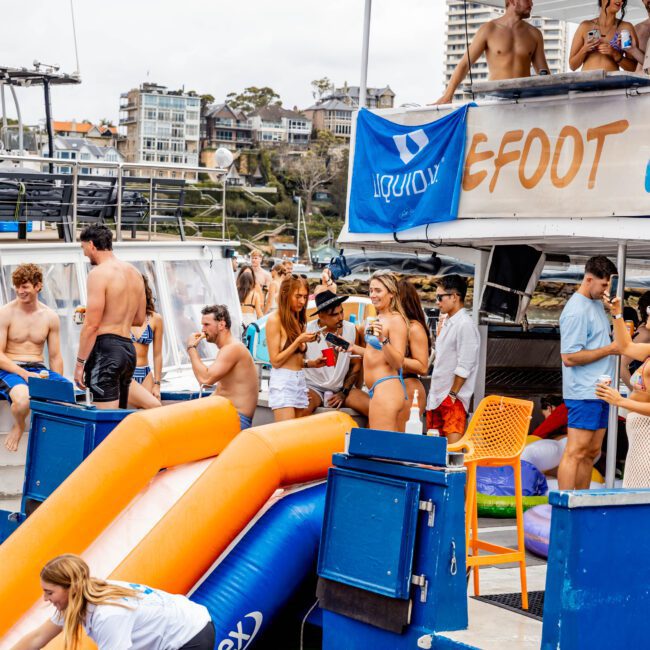 A lively boat party scene with people in swimsuits enjoying themselves. Some are on an inflatable slide leading into the water, while others chat on the boat. The background features buildings and a cloudy sky.