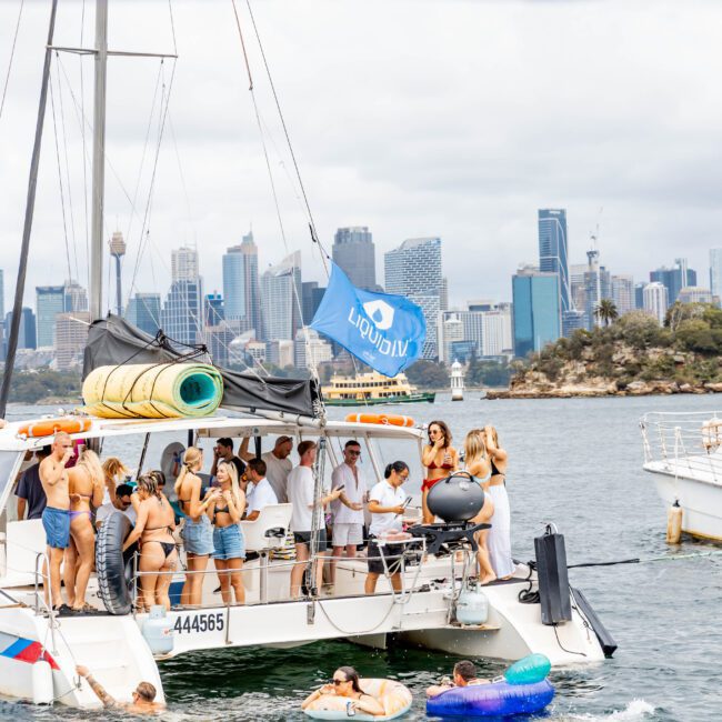 A group of people in swimwear are socializing on a catamaran in a bustling harbor. Inflatable toys float nearby, and the city skyline is visible in the background under a cloudy sky.
