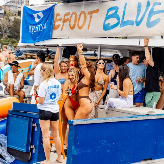 A group of people enjoying a boat party. Several individuals are dancing and waving, while others stand around talking. A banner reads "EFFORT BLUE" and people are wearing casual summer attire. A few are holding drinks.