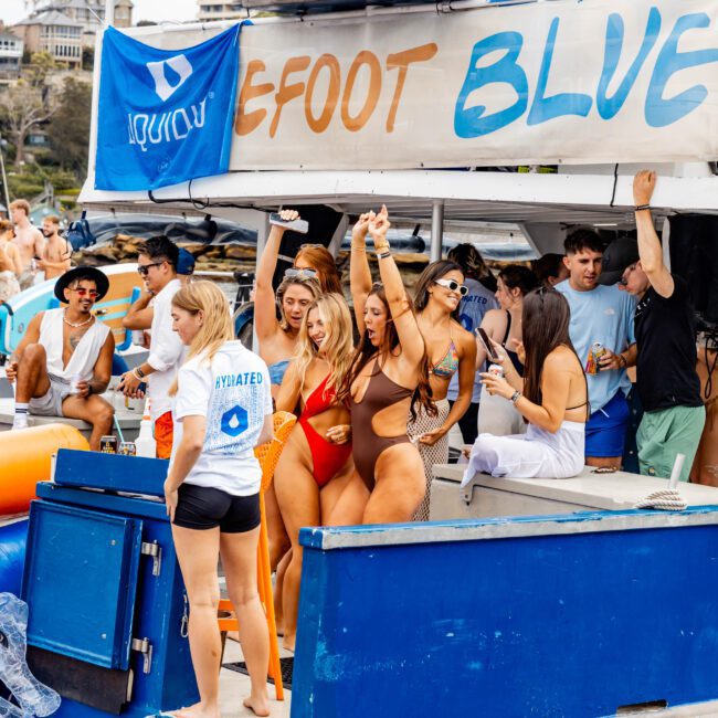 A lively group of people are enjoying a party on a boat. Several women in swimsuits dance and raise their hands, while others stand and chat. A banner that reads "6FOOT BLUE" is visible. The background shows buildings and greenery.