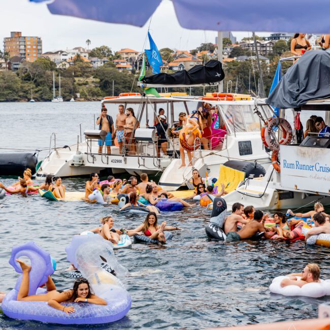 People are enjoying a party on a river, surrounded by boats and inflatables. Some are swimming, while others relax on floating devices. The scene is lively, with a mix of colorful inflatables and boats in the background.