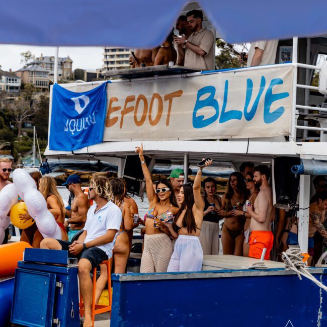 A lively group of people enjoying a boat party, some standing and some sitting. A banner reads "Barefoot Blue." The background shows buildings and more watercraft.