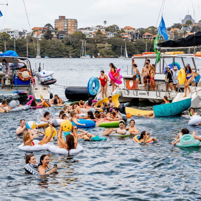 A lively scene on the water with many people on colorful inflatables, including swans and unicorns, near several boats. The sky is overcast, and houses are visible on the distant shore. People are enjoying the festive atmosphere.