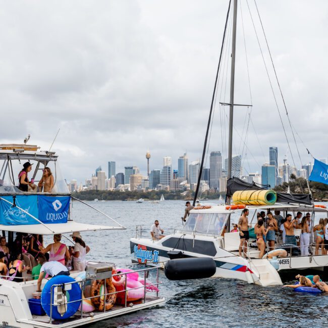 Boats filled with people partying on the water, with a city skyline visible in the background. Many people are on the decks enjoying the event, while others are in the water on floats. The sky is cloudy, adding a dramatic backdrop.