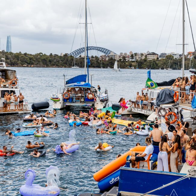 A lively party scene on the water with people on inflatable floats surrounding several boats. Attendees are enjoying the event under a cloudy sky, with a city skyline visible in the background.