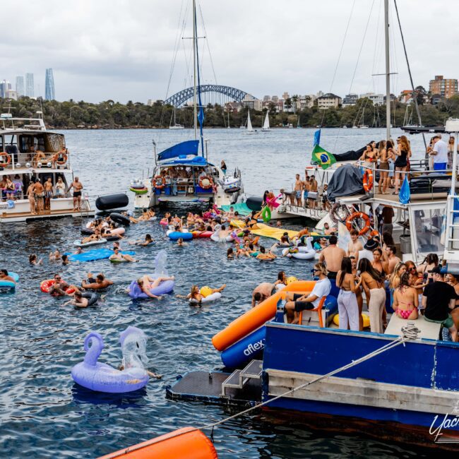 A lively scene on the water with people on boats and colorful inflatable floats in a harbor. The Sydney city skyline and the Harbour Bridge are visible in the background under a cloudy sky.