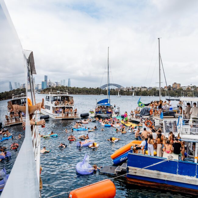 A lively scene on a harbor features multiple boats filled with people partying. Many individuals are swimming or floating on colorful inflatables in the water. The background shows a city skyline and an iconic bridge. The atmosphere is festive and cheerful.