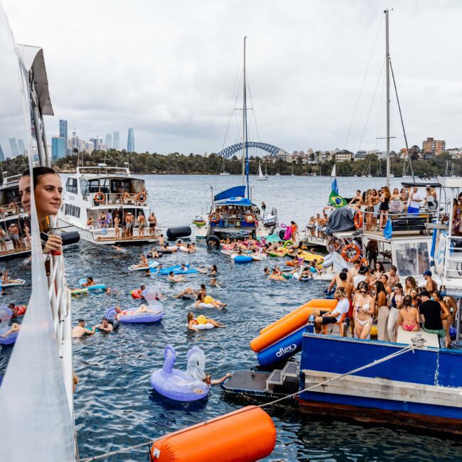 People enjoying a lively yacht party on a river, with inflatables in the water and several boats anchored nearby. A person leans out from a boat on the left side. The background features a cityscape with a bridge and tall buildings.