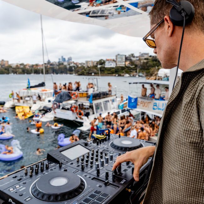DJ on a boat using a mixer, wearing headphones. A lively crowd on inflatables and other boats can be seen in the water. The reflection of the scene is visible on the ceiling. A beer bottle is near the mixer. A "Yacht Social Club" logo is in the corner.