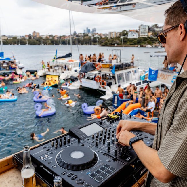 DJ playing music on a boat, overlooking a lively pool party. People are floating on inflatables and swimming. The backdrop features a city skyline and a bridge. Sunny weather and festive atmosphere with drinks on the DJ deck.